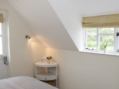 A bedroom corner with a white bedside table holding a clock and a small plant under a wall light and a window with a yellow patterned roman blind at Bicton Lodge in Clun