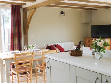 A dining area with a wooden table and chairs next to a kitchen island with flowers and bottles at Bicton Lodge in Clun