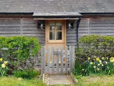 A wooden front door with a small pitched roof and gate surrounded by bushes and flowers at Bicton Lodge in Clun