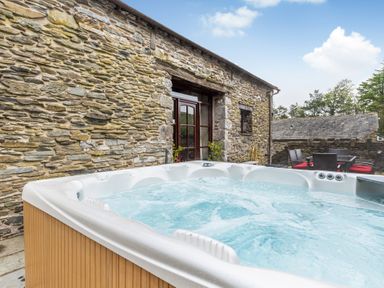 An outdoor hot tub on a stone patio next to a stone building with wooden doors and outdoor dining furniture with red cushions at Hill Side Barn in Pennington near Ulverston