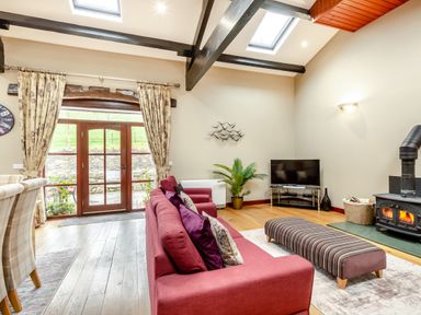 A living room with a red sofa, striped ottoman, television, wood stove, and glass doors at Hill Side Barn Pennington near Ulverston