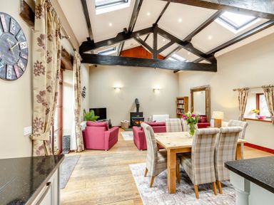 A dining and living area with wooden beams a wood stove pink sofas and a wooden dining table with plaid chairs at Hill Side Barn in Pennington near Ulverston