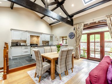 A dining area with a wooden table and six plaid chairs next to a kitchen with white cabinets and a black countertop at Hill Side Barn in Pennington near Ulverston