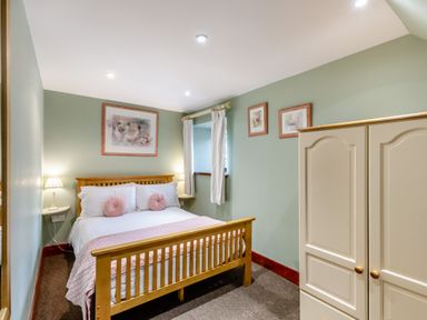 A bedroom with a wooden bed with white and pink bedding two bedside tables with lamps a window with white curtains and a cream wardrobe at Hill Side Barn in Pennington near Ulverston