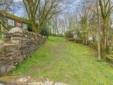 A grassy path bordered by a stone wall and trees with a small green building labeled Daisy Cottage at Hill Side Barn in Pennington near Ulverston