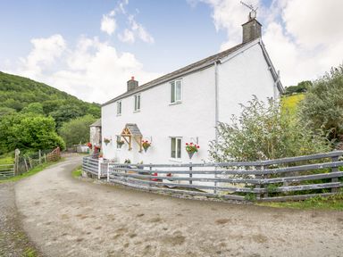 A white cottage with flower baskets on the wall and a grey fence along a curved dirt road at Ty Nant in Betws-y-Coed