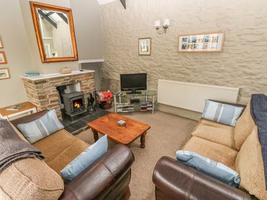 A living room with two sofas a wooden coffee table a fireplace with a wood stove a television on a glass stand and framed pictures on a stone wall at Foxglove Cottage in Little Haven near Haverfordwest