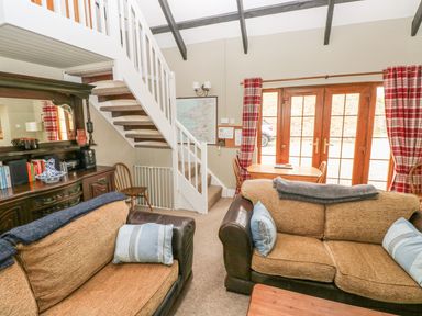 A living room with two brown sofas a wooden dining table and chairs a staircase with white railing and glass doors with red plaid curtains at Foxglove Cottage Little Haven near Haverfordwest