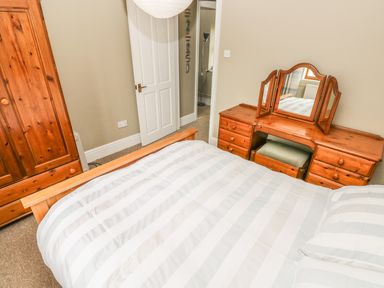 A bedroom with a wooden dresser with a three-panel mirror a wooden wardrobe and a bed with striped bedding at Foxglove Cottage in Little Haven near Haverfordwest