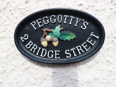 A black oval house sign with white letters reading Peggotty's 2 Bridge Street with a design of three acorns and two oak leaves at Peggotty's in Kelsale near Saxmundham