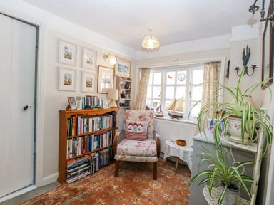 A small sitting room with a floral armchair near a window with model sailboats on the sill next to a wooden bookshelf and plants on a metal stand at Peggotty's in Kelsale near Saxmundham