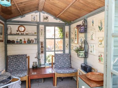 A wooden garden shed with two cushioned chairs a wooden table shelves with crockery and plants at Peggotty's in Kelsale near Saxmundham