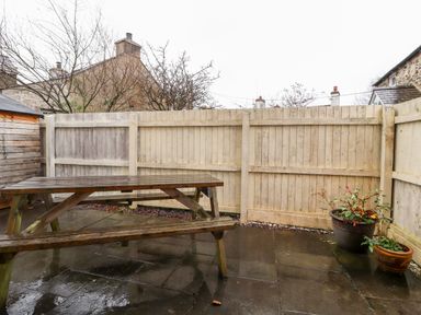 A wooden picnic table on a wet paved patio with a wooden fence and two potted plants at Bryn Seion in Y Felinheli