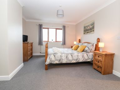 A bedroom with a wooden bed with patterned bedding and yellow pillows two wooden bedside tables with lamps a wooden dresser with a television and a window with grey curtains at Bryn Seion in Y Felinheli