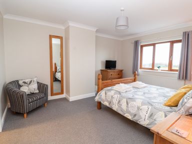 A bedroom with a double bed and patterned bedding a wooden dresser with a television a striped armchair and a wall mirror at Bryn Seion in Y Felinheli