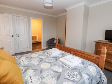 A bedroom with a wooden bed, folded towels on the bed, a striped armchair, a wooden dresser, and an open door leading to a bathroom at Bryn Seion in Y Felinheli
