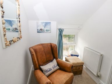 A small room with a brown leather armchair a beige ottoman a window with blue curtains and a wall mirror with shells at Hillbrook House in Nolton Haven