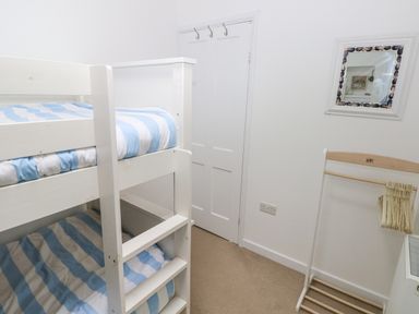A small bedroom with a white bunk bed with blue and white striped bedding a door with hooks a wooden clothes rack with hangers and a wall mirror at Hillbrook House in Nolton Haven