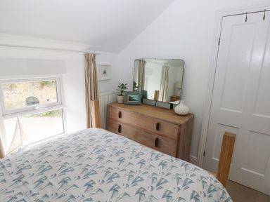A bedroom with a wooden bed with patterned bedding a wooden dresser with a mirror potted plant and picture frame near a window at Hillbrook House in Nolton Haven
