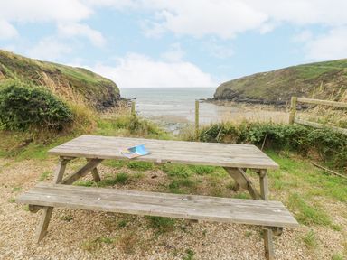 A wooden picnic table on a gravel patch overlooking a small beach and ocean between grassy cliffs at Hillbrook House in Nolton Haven