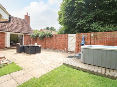 A backyard with a patio table and chairs a hot tub on a wooden platform a brick wall and an open door leading into a house at Oak Tree Lodge in Crostwick near Norwich