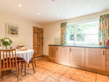 A kitchen with wooden cabinets a window with patterned curtains and a dining table with chairs at Dorothy's Cottage in Alnmouth