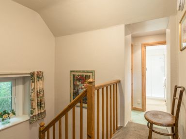 A staircase hallway with wooden banister, a chair, a small window with floral curtains, and a view into a bathroom at Dorothys Cottage in Alnmouth