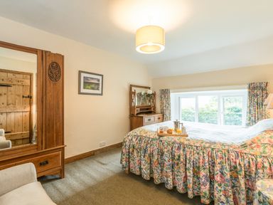 A bedroom with a floral bedspread on a bed a wooden wardrobe with a mirror a wooden dresser with a mirror and a window with floral curtains at Dorothy's Cottage in Alnmouth