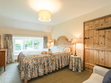 A bedroom with a wooden bed and floral bedspread floral curtains wooden door and bedside lamp at Dorothy's Cottage in Alnmouth