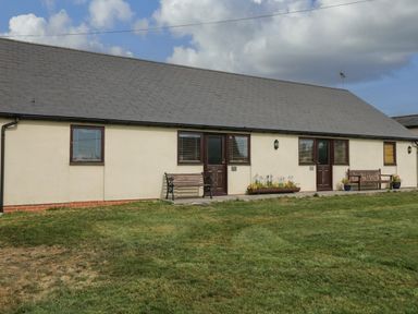 The exterior of a single story building with grass in front and benches near the doors at Brindle Cottage in Royal Wootton Bassett