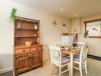 A dining area with a wooden table and four chairs with cushions a wooden cabinet with shelves fruit bowl and plants at Brindle Cottage in Royal Wootton Bassett