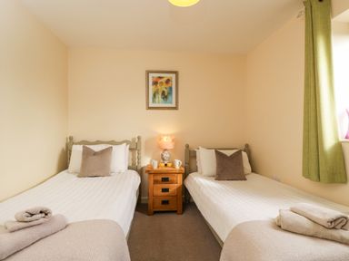 A bedroom with two single beds, a wooden nightstand with a lamp, cups, and orange juice, and a window with green curtains at Brindle Cottage Royal Wootton Bassett