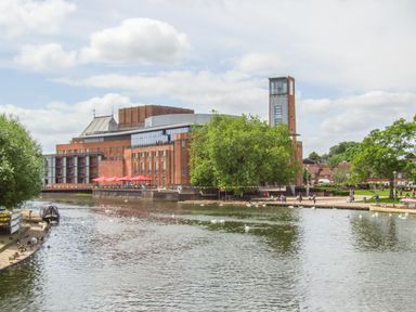 A river with ducks and people walking next to a large brick building at Highcroft in Stratford-Upon-Avon