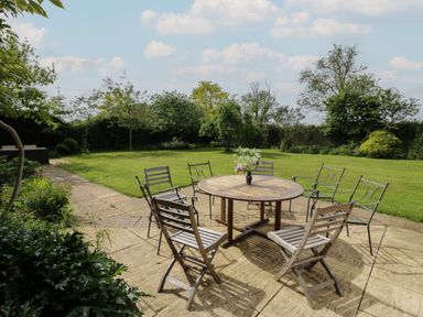 An outdoor patio with a round wooden table and six chairs on a stone tiled surface with a garden and trees in the background at Highcroft  in Stratford-Upon-Avon
