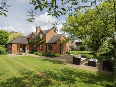 A brick house with outdoor seating areas on a patio and a lawn with trees at Highcroft in Stratford-Upon-Avon