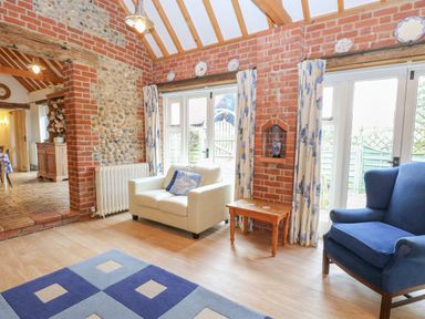A sitting room with brick walls a white armchair a blue armchair a wooden side table and a blue patterned rug at Stable Cottage in Necton near Swaffham
