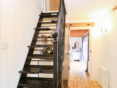 A hallway with black metal stairs and a door leading to a room with wooden beams a white dresser and a purple lamp at Stable Cottage in Necton near Swaffham
