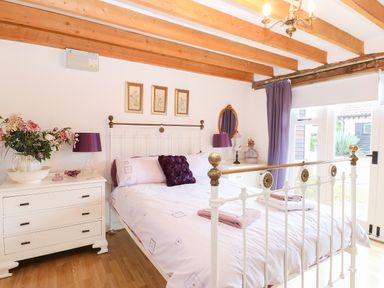 A bedroom with a white metal bed frame purple cushions bedside tables with lamps flowers and curtains at Stable Cottage in Necton near Swaffham