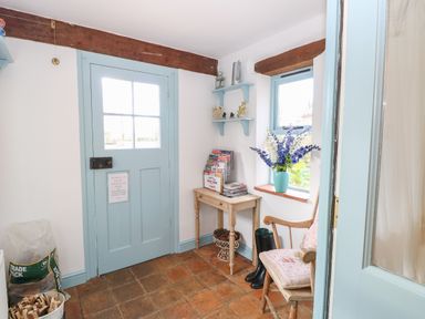 A small room with a blue door a wooden table with magazines and a vase of flowers a wooden chair with cushions and a pair of black boots at Stable Cottage in Necton near Swaffham