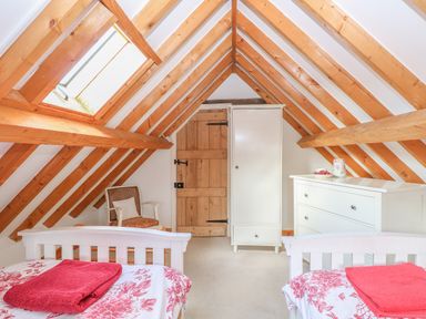 An attic bedroom with wooden beams two single beds with red and white bedding a wooden door a white wardrobe a white chest of drawers and a chair by a skylight window at Stable Cottage in Necton near Swaffham