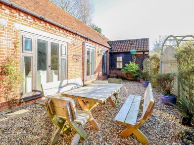 An outdoor patio area with wooden table and benches surrounded by plants and a brick house at Stable Cottage Necton near Swaffham