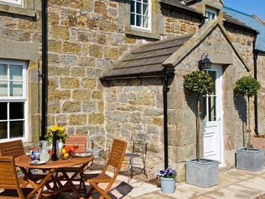 An outdoor patio with a wooden table and chairs set against a stone house wall at The Old Farmhouse in Lowick