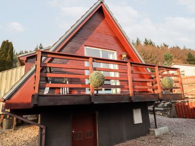 A red wooden chalet with a balcony and hanging plants in a wooded area at Larchfield Chalet 2 in Heights of Achterneed near Strathpeffer