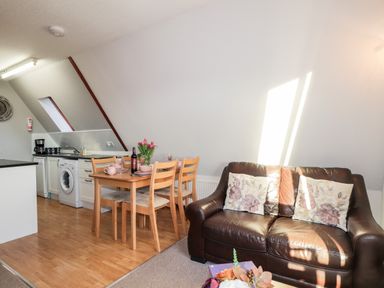 A living area with a brown leather sofa with floral cushions a wooden dining table with four chairs and a kitchen area with appliances at Larchfield Chalet 2 Heights of Achterneed near Strathpeffer