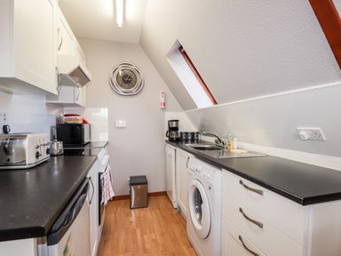 A galley kitchen with black countertops white cabinets a washing machine and a wall clock at Larchfield Chalet 2 Heights of Achterneed near Strathpeffer