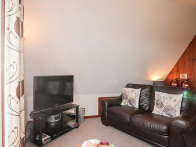 A living room with a black leather sofa with floral cushions and a television on a black stand at Larchfield Chalet 2 Heights of Achterneed near Strathpeffer