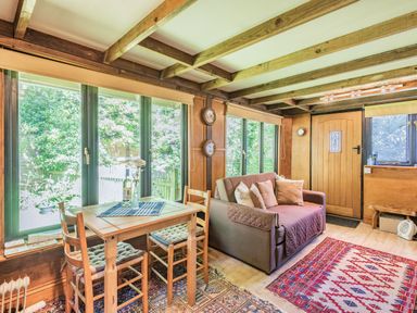 A sitting room with a small wooden dining table and chairs beside large windows and a sofa with cushions at Endymion in Linwood near Ringwood New Forest
