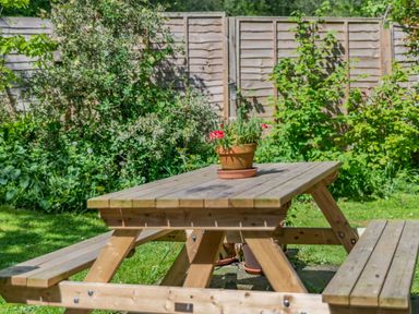 A wooden picnic table with a flower pot on top in a garden with green plants and a wooden fence at Endymion in Linwood near Ringwood New Forest
