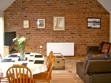 A dining area with a round table and wooden chairs and a living space with leather sofas against a brick wall at Happy Union Stables in Abbeycwmhir near Rhayader