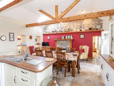 A dining area with a wooden table and chairs next to a kitchen island in a room with exposed wooden beams and a stone fireplace at Prague House in Lettermore County Galway
